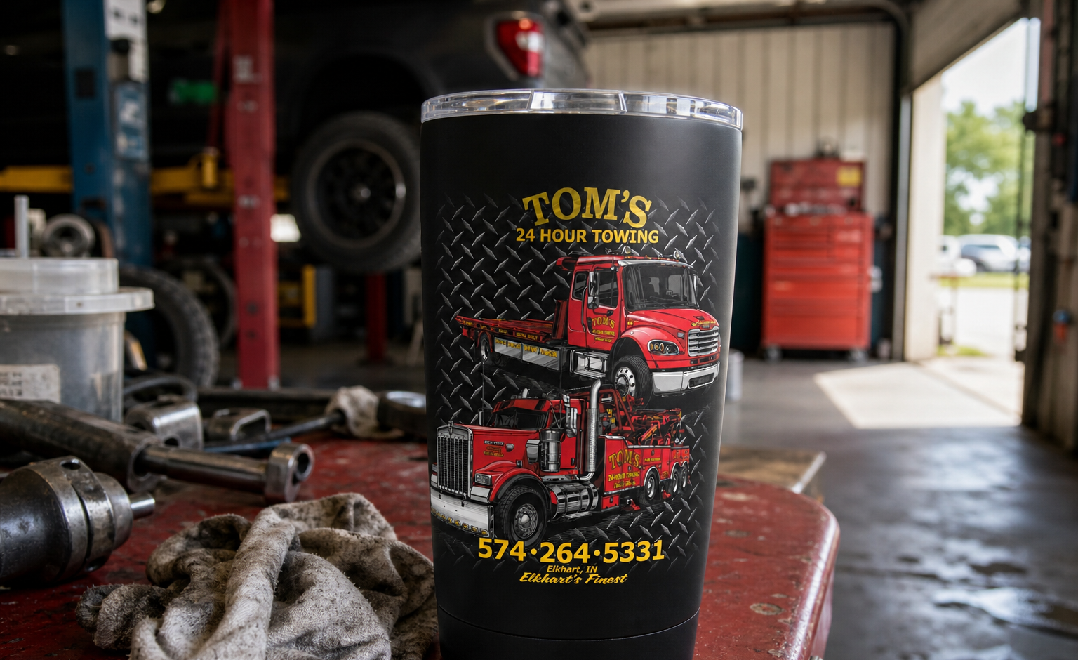 Matte black 20 oz tumbler with full-color Tom's 24 Hour Towing truck artwork, sitting on a workbench in a towing shop.
