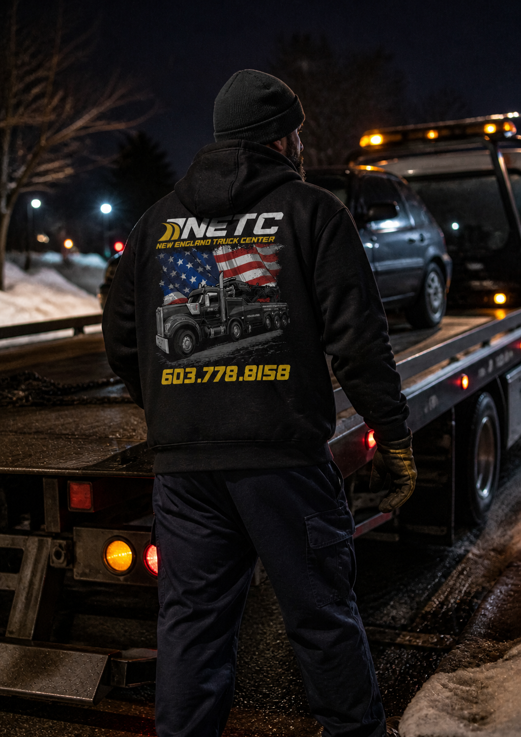Black hoodie featuring New England Truck Center towing artwork, worn beside a tow truck at night.
